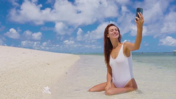 Hermosa mujer con el pelo que sopla usando el teléfono inteligente y haciendo selfie en el fondo del Océano Índico — Vídeos de Stock