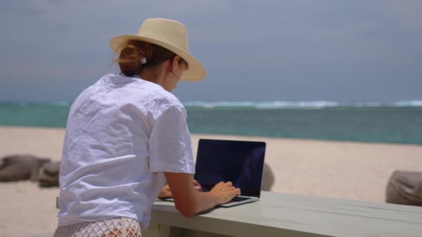 Business woman in yellow hat using laptop computer on the background of a tropical beach with turquoise water. Vacation, freelance job, leisure activity concept — Stock Video