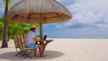 Serbest çalışma, seyahat ve tatil konsepti. Sarı şapkalı bir adam okyanusun kumsalında oturuyor. Denizde tatildeyken çalışmak