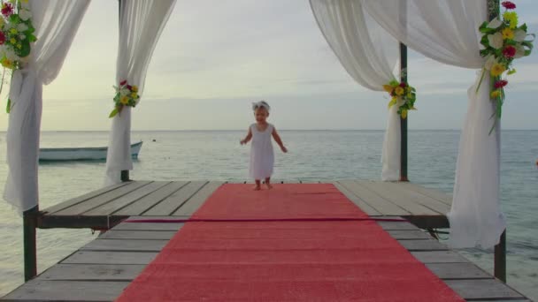 Маленькая двухлетняя девочка в белом платье прогуливается по цветочному пирсу на закате на берегу Индийского океана. Концепция свадебного видео — стоковое видео