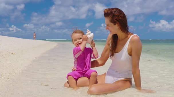 Ibu dan putrinya bermain di air jernih di pantai Samudera Hindia. Ibu dan bayi memegang cangkang besar di tangan mereka. Suara laut. — Stok Video