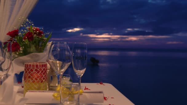 Decorar una mesa festiva. Decoración de mesa de boda. Mesa de noche romántica para dos personas en la playa al atardecer en el fondo de hermosas nubes — Vídeo de stock