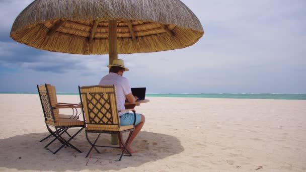 Concepto independiente, viajes y vacaciones. Un hombre con sombrero amarillo sentado en el fondo de la playa del océano. Trabajar de vacaciones en el mar — Vídeos de Stock