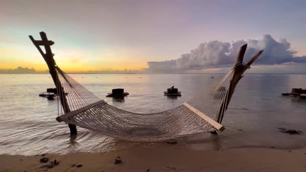 Gün batımı. Sakin dalgalar okyanustan kıyıya ulaşırken rüzgarın yanında uzanan boş bir hamağın statik görüntüsü. Hint Okyanusu. Mauritius. — Stok video