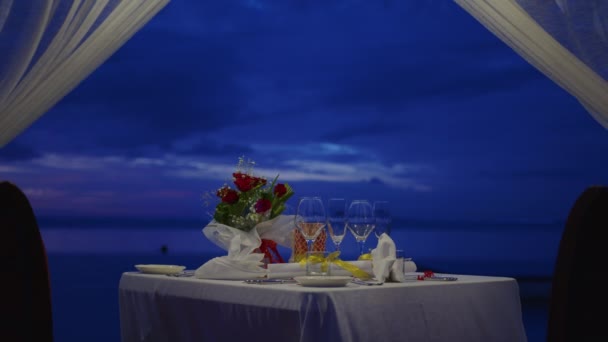 Decorar una mesa festiva. Decoración de mesa de boda. Mesa de noche romántica para dos personas en la playa al atardecer en el fondo de hermosas nubes — Vídeos de Stock