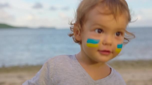 Zamknij portret małego dziecka z flagą Ukrainy na twarzy siedzi sam. Protest przeciwko rosyjskiej inwazji wojennej na Ukrainie. Flaga ukraińska, Koncepcja protestujących — Wideo stockowe
