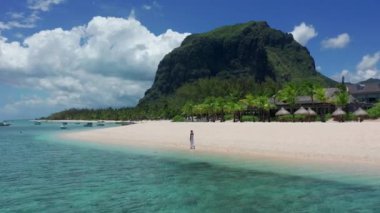 Tropikal tatil beldesi kıyı turizmi kavramı. Beyaz elbiseli genç bir kadın tropik sahilde yürüyor. Mauritius sahilinde ayakları suda yürüyen bir kızın geniş açılı görüntüsü.