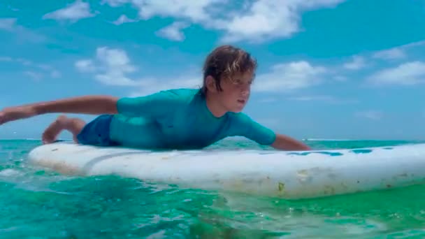 Surfare paddlar sina händer på en surfbräda. Pojke surfare väntar vågen i havet på solig tropisk dag med gröna kullar vid horisonten — Stockvideo