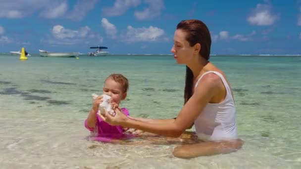 Mutter und Tochter spielen im klaren Wasser am Ufer des Indischen Ozeans. Mutter und Baby halten eine große Muschel in den Händen. Das Geräusch des Ozeans — Stockvideo