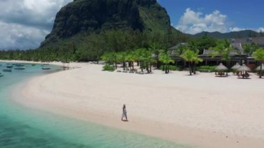 Tropikal tatil beldesi kıyı turizmi kavramı. Beyaz elbiseli genç bir kadın tropik sahilde yürüyor. Mauritius sahilinde ayakları suda yürüyen bir kızın geniş açılı görüntüsü.
