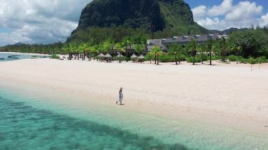 Tropikal tatil beldesi kıyı turizmi kavramı. Beyaz elbiseli genç bir kadın tropik sahilde yürüyor. Mauritius sahilinde ayakları suda yürüyen bir kızın geniş açılı görüntüsü.