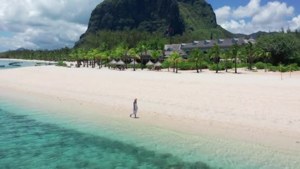 Koncepció trópusi üdülőhely partmenti turizmus. Egy fehér ruhás fiatal nő sétál a trópusi parton. Széles kép egy lányról, aki Mauritius tengerpartján sétál a lábával a vízben. — Stock videók