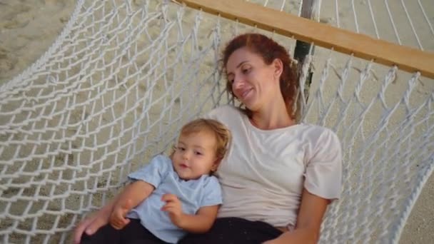 Fetița îmbrățișează ușor mama într-un hamac într-un stoc de mișcare lentă hamac. Bebelușul se odihnește cu o mamă fericită într-un hamac pe ocean. Conceptul de copii, copil, părinte, copilărie, maternitate — Videoclip de stoc