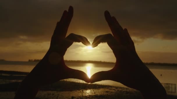 Glad tjej på stranden vid solnedgången. Bild gjord med fingrar. Händer flicka form av hjärta. Sommardröm. Frihetens lycka vid solnedgången. Solljus mellan fingrarna. Silhuett av lycklig flicka på havet — Stockvideo