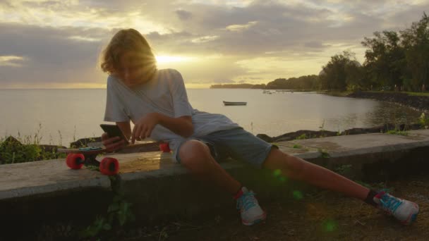 Sevimli bir genç güzel bir günbatımının arka planında oturarak cep telefonunu kullanır. Kaykaycı egzersizden sonra dinleniyor — Stok video