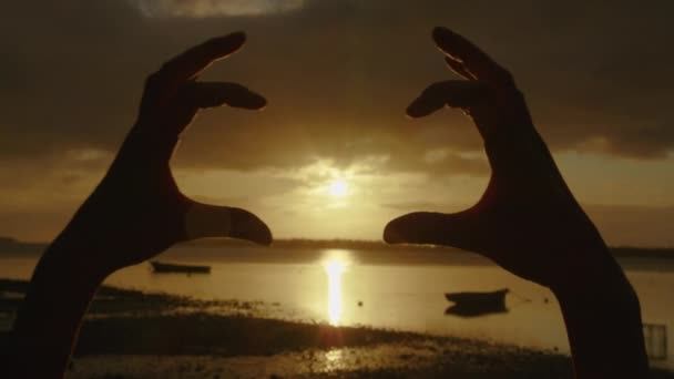 Glad tjej på stranden vid solnedgången. Bild gjord med fingrar. Händer flicka form av hjärta. Sommardröm. Frihetens lycka vid solnedgången. Solljus mellan fingrarna. Silhuett av lycklig flicka på havet — Stockvideo