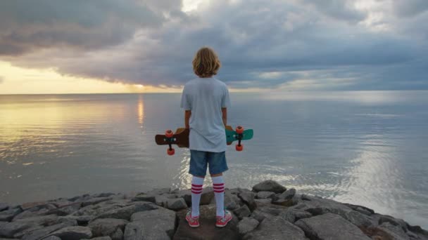 Niño con un longboard en una hermosa playa. Adolescente skateboarding en fondo puesta de sol — Vídeos de Stock