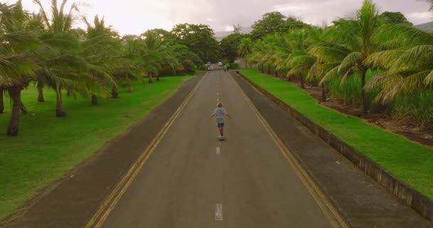 Підліток їде на дошці вздовж красивої дороги з зеленими пальмами — стокове відео