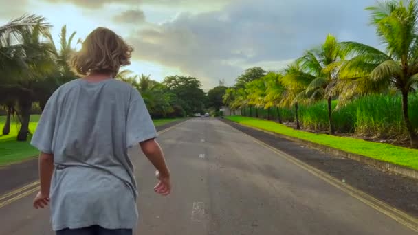 Un adolescent monte un longboard le long d'une belle route avec des palmiers verts — Video