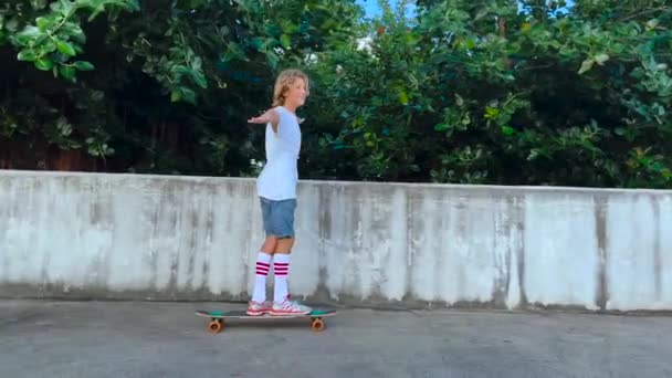 Cheerful little boy with long wavy hair in rides on a long skateboard. Learns to do tricks. Gray concrete wall in the background — Stockvideo