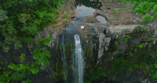 Woman in white with arms raised on top of beautiful waterfall looking at Sunrise. Aerial view of high waterfall in green jungle — Video