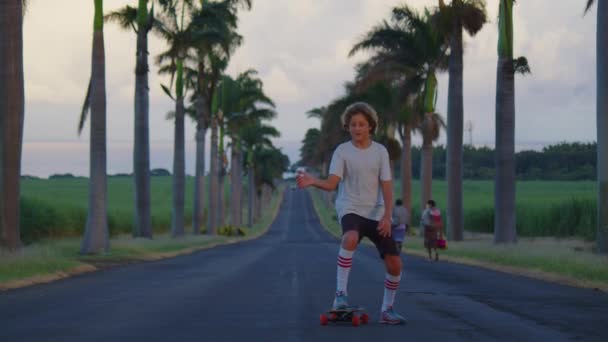 Un adolescente con i capelli lunghi cavalca uno skateboard lungo una bella strada con palme. Los Angeles. Ragazzo longboarder cavalcando il suo lungo bordo giù per la montagna in bella natura panoramica su strada — Video Stock