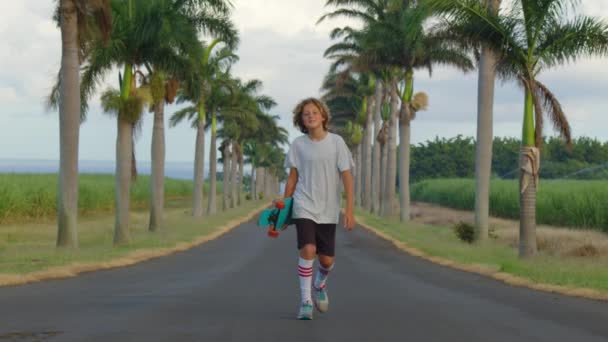 Un adolescent aux cheveux longs fait du skateboard le long d'une belle route avec des palmiers. Los Angeles. Garçon longboarder chevauchant sa longue planche en bas de la montagne dans la belle nature pittoresque sur la route — Video