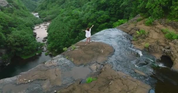 Woman in white with arms raised on top of beautiful waterfall looking at Sunrise. Aerial view of high waterfall in green jungle — Vídeo de Stock