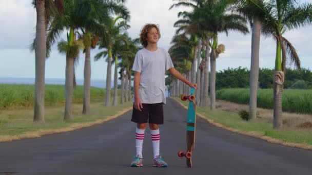 Jonge coole man, tiener hipster in Californië rijdt op zijn skateboard op het strand. Actie breed schot van de mens uit te voeren skateboard trucs op zonnige dag in toeristische plek. Weg met palmbomen — Stockvideo