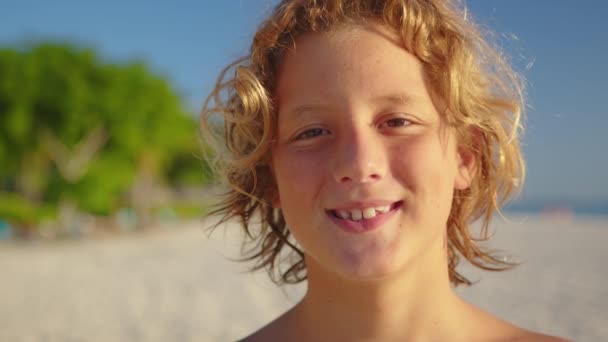Egy fiatal fiú portréja, hosszú hajjal, ahogy mosolyog a kamerába. Közelkép. Kék ég van a háttérben.. — Stock videók