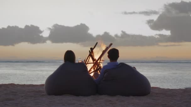 Happy couple resting in front of burning yellow campfire at evening on the coastline. Beautiful couple resting by fire, young loving couple hugs and dreams enjoying romantic weekend near campfire — Stockvideo