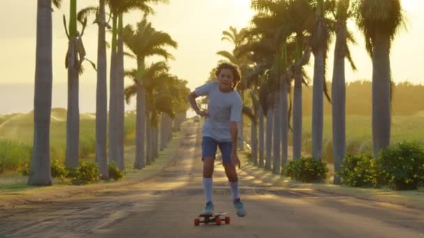 Happy care free boy skateboarding down street at sunset with hands up in air. Palm trees by a blue sky. Driving through the sunny Beverly Hills. Los Angeles, California. — стоковое видео