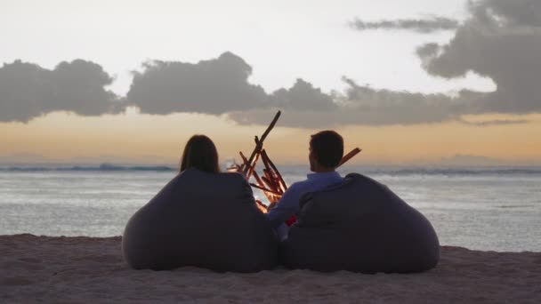 A young couple is sitting on the beach by the fire. The concept of love and friendship in nature. Back view — стоковое видео