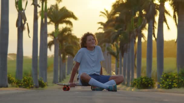 Um adolescente está patinando ao longo de uma bela estrada com palmeiras. Silhueta de um menino com um skate — Vídeo de Stock