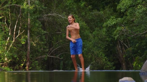 The boy dances cheerfully in the park against the backdrop of green trees. A beautiful reflection on the surface of the water of a dancing teenager. Water splash — Stockvideo