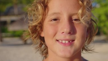 Kameraya gülümseyen uzun saçlı bir çocuğun portresi. Yakın plan. Arka planda mavi bir gökyüzü var..