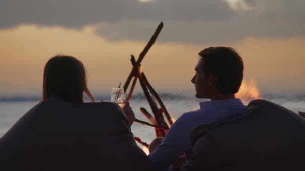 Pasangan muda sedang duduk di pantai dekat api. Konsep cinta dan persahabatan di alam. Tampilan belakang — Stok Video