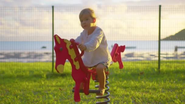 Baby rider på en häst gunga på lekplatsen. Lycklig familj, barn leker på en gunga på våren lekplats, barn utomhus aktiviteter, livet för små människor på helgerna, barn dröm — Stockvideo