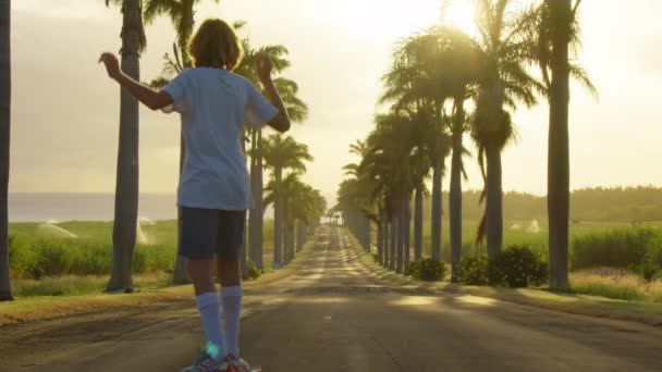 Happy care free boy skateboarding down street at sunset with hands up in air. Palm trees by a blue sky. Driving through the sunny Beverly Hills. Los Angeles, California. — стоковое видео