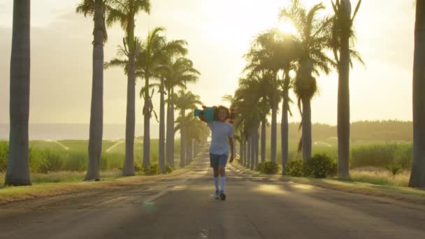 A teenager is skating along a beautiful road with palm trees. Silhouette of a boy with a skateboard — Stockvideo