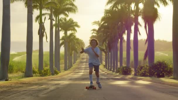 Happy care free boy skateboarding down street at sunset with hands up in air. Palm trees by a blue sky. Driving through the sunny Beverly Hills. Los Angeles, California. — стоковое видео