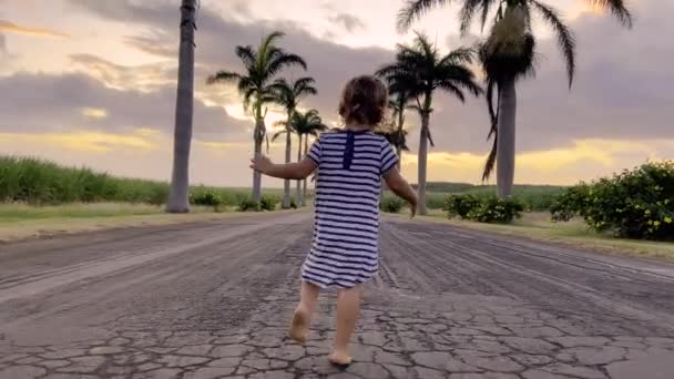 Ένα κοριτσάκι με ριγέ φόρεμα τρέχει στο δρόμο με φοίνικες στο φόντο ενός όμορφου ηλιοβασιλέματος. — Αρχείο Βίντεο