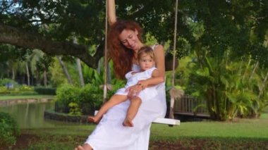 Beyaz elbiseli anne ve kızı haftasonunu yeşil bir parkta salıncakla geçiriyorlar. Doğada aile tatili. Bir çocuğa annelik bakımı