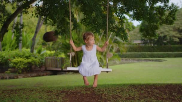 하얀 드레스를 입은 한 어린 소녀 가 공원에서 밧줄 그네를 타고 있습니다. 옥외 오락의 개념 — 비디오