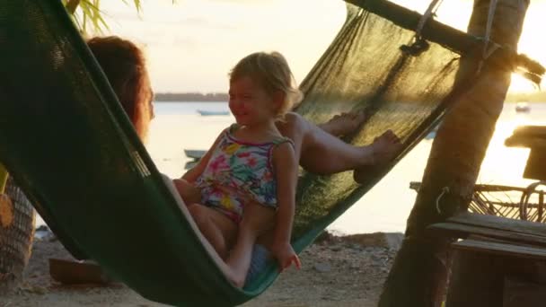 若い母親は ビーチでのハンモックで娘と一緒に座って笑って 日没の背景に笑顔 日没時に熱帯ハンモックでリラックスした幸せな家族 夏の贅沢休暇 — ストック動画