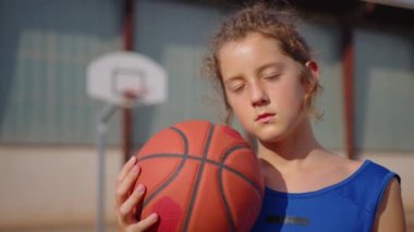Joyful young sportsman posing with ball in sunlight on outdoor court. Portrait of smiling caucasian man standing at the background of basketball hoop and looking at camera. High quality 4k footage