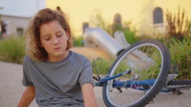 The boy sits on the road after falling off a bicycle, calms the pain in his elbow. Broken bike in the background. High quality 4k footage