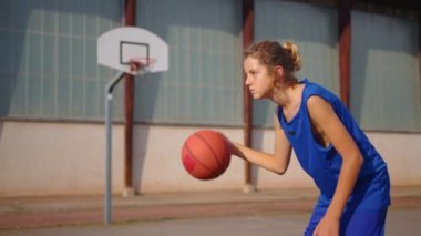 Active male basketball player practicing ball handling skill, dribbling ball between legs on outdoor court in rays of early morning rising sun during streetball training in neighborhood.