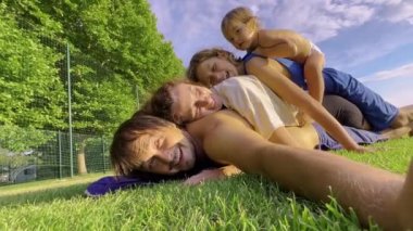 A happy family of four is playing on the green grass. Cute children climbed on their parents laughing and playing. Friendly family in the park. High quality 4k footage