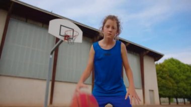 Active male basketball player practicing ball handling skill, dribbling ball between legs on outdoor court in rays of early morning rising sun during streetball training in neighborhood.